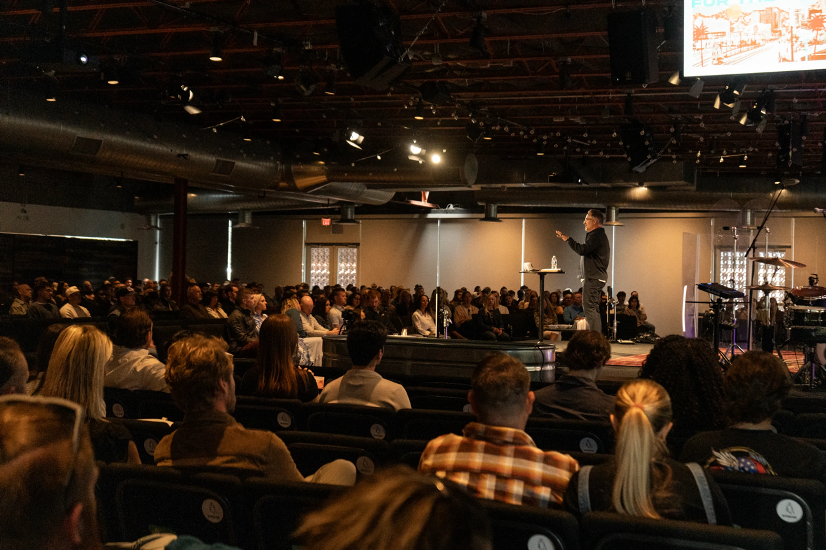A pastor stands on a brightly lit stage, speaking to a large congregation seated in a modern auditorium. The photo is taken from the back of the room.